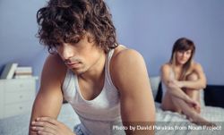 Upset man sitting on edge of bed after argument with woman in background 5ngPaM
