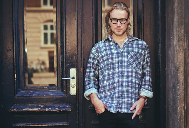 Man in glasses with long blonde hair leaning against a wooden door outside