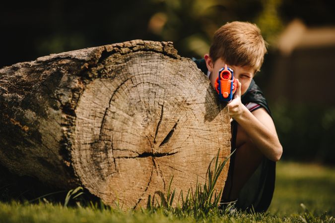 Young child with toy gun hiding behind a log outdoors in park
