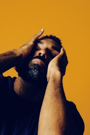 Tired Black man in yellow studio with both hands to his face