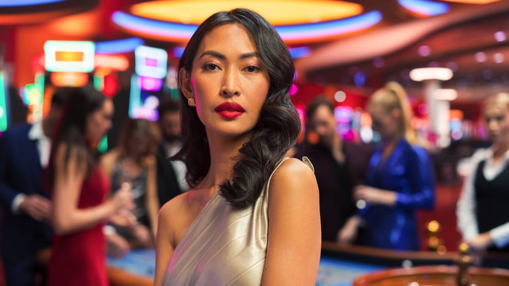 In a Modern Casino Building: Asian Woman Posing Confidently, Looking at Camera, Wearing a Fashionable Grey Dress.