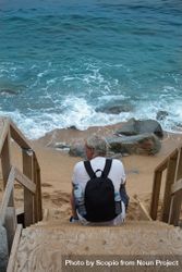 Back view of middle aged man with backpack sitting on wooden stairs by the sea bEJL10
