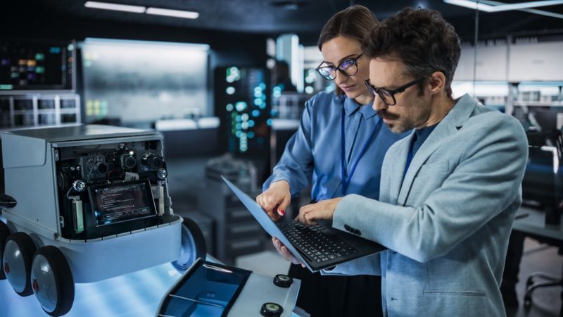 Portrait of Male and Female Engineers Developing a Remotely Controlled Robot in an Industrial Research Facility. Specialists Using Laptop Computer, Working on Machine Improvement, Brainstorming