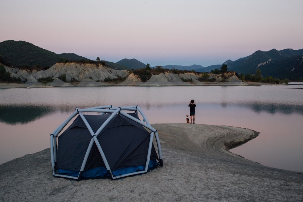 Man with binoculars and dog looking over lake with tent at sunset