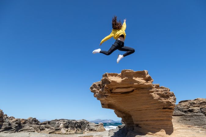 Sportive woman jumping above rock against blue sky