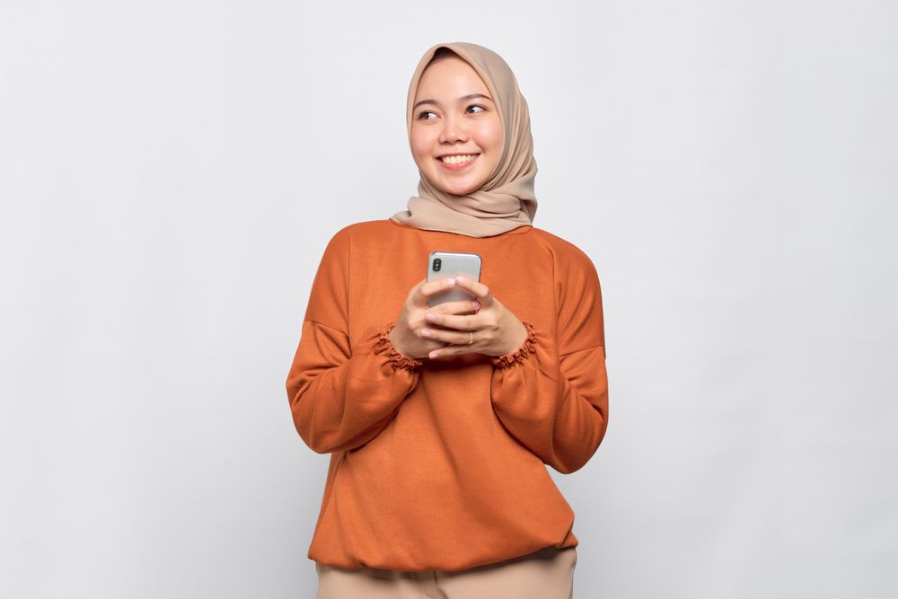 Smiling Muslim woman in headscarf and orange shirt with phone and looking away