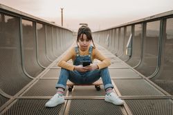 Girl with smartphone sitting on skateboard on walkway