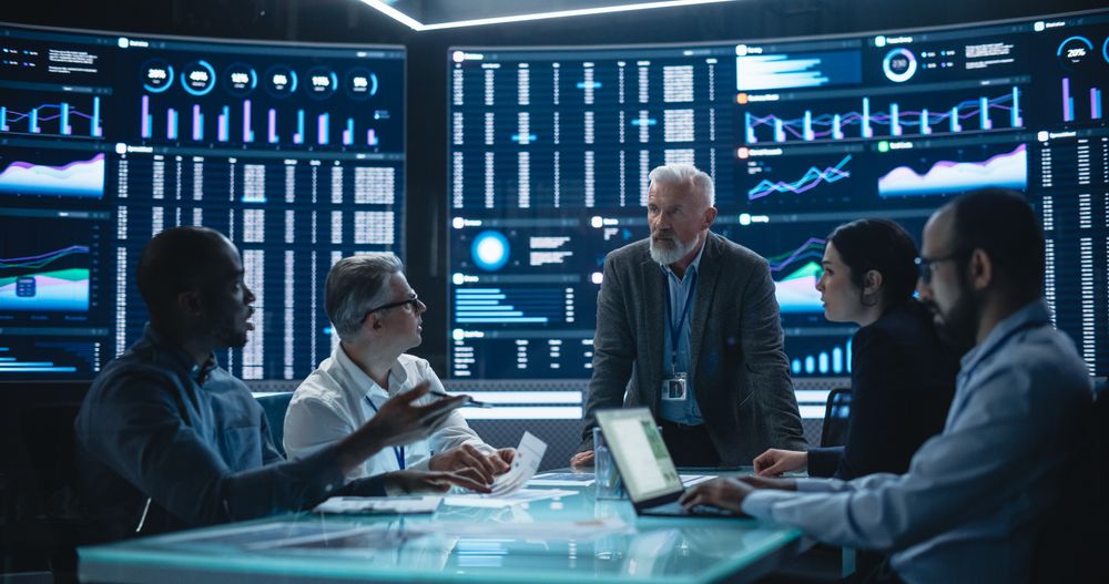 Team Of Data Scientists and Software Developers Discussing Reports in Monitoring Room with Analytics Feed on Big Digital Screen.