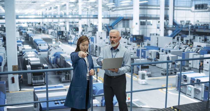 Mature Engineer and Young Japanese Technician Having a Conversation and Using Laptop Computer. Specialists Monitoring, Analyzing Conditions at a Modern Electronics Factory with Automated Robots