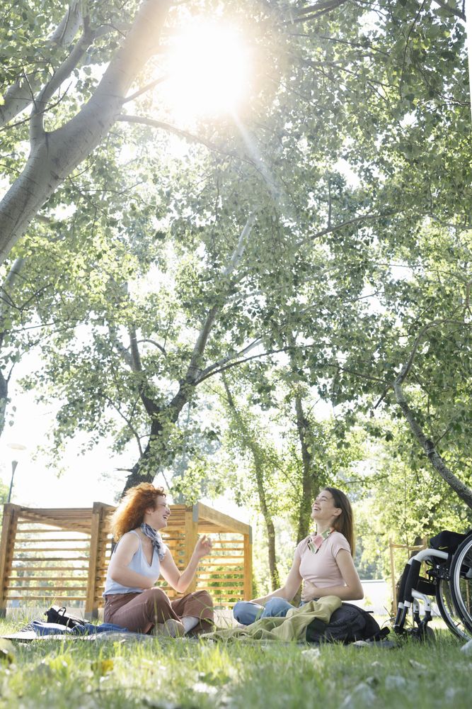 Woman with a disability sitting next to her wheelchair having a picnic with friend, vertical