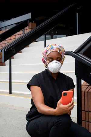 Nurse in N95 mask and scrubs checks cell phone while sitting outside on steps