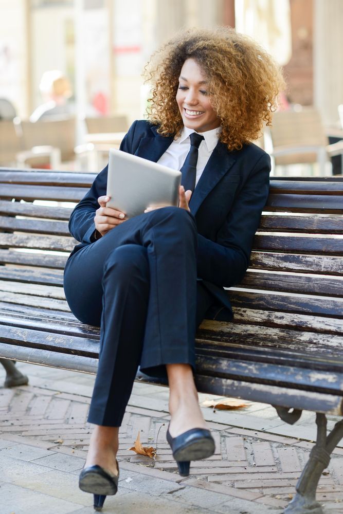 Woman in suit laughing at something on her digital tablet on outdoor bench