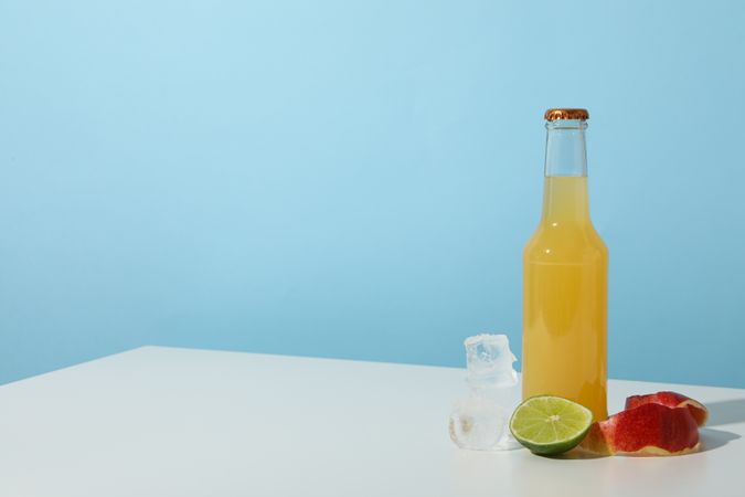Bottle with apple cider, piece of lime, ice cubes and apple peels on blue background, space for text
