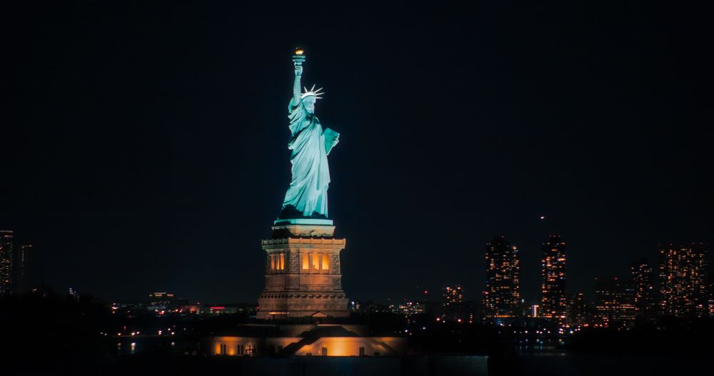 Aerial View of the Illuminated Statue of Liberty. Iconic Travel Destination and Tourist Attraction with Manhattan and Brooklyn Office Buildings All Lit Up at Night. Nightlife in New York City