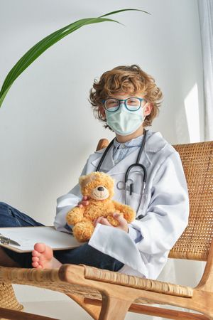 Boy doctor with toy bear on chair on white background