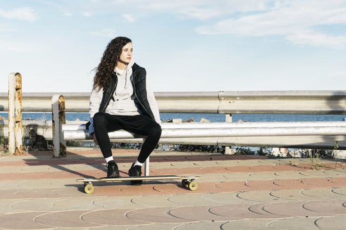 Teenage girl sitting on a bench with her feet on the longboard in front of the sea