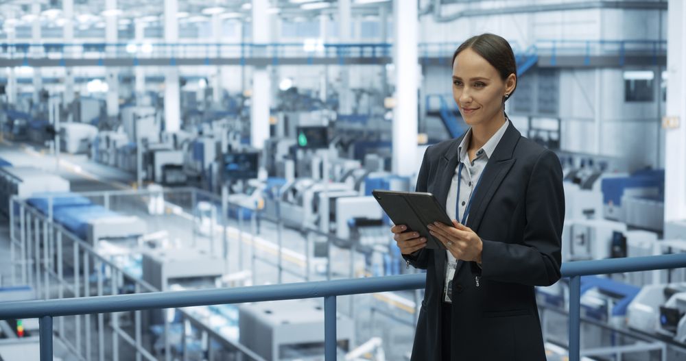 Portrait of a Happy Engineer Using Laptop Computer and Looking Around a Factory Facility with Automated Robotic Arms Producing Modern Electronic Components for Different Industries