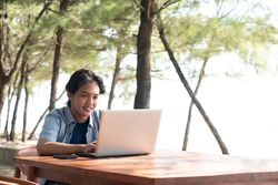 Happy Asian Young businessman work at outdoor with laptop. Concept of remote and freelance work. Young successful man wearing casual blue cloth.