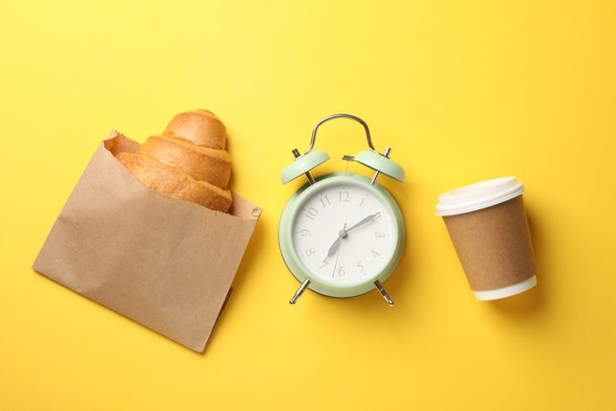 Top view of croissant, clock and takeaway coffee on yellow background, space for text