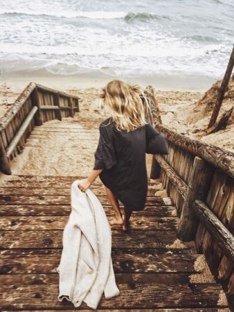 Back view of blonde woman holding a coat getting down wooden steps by seashore