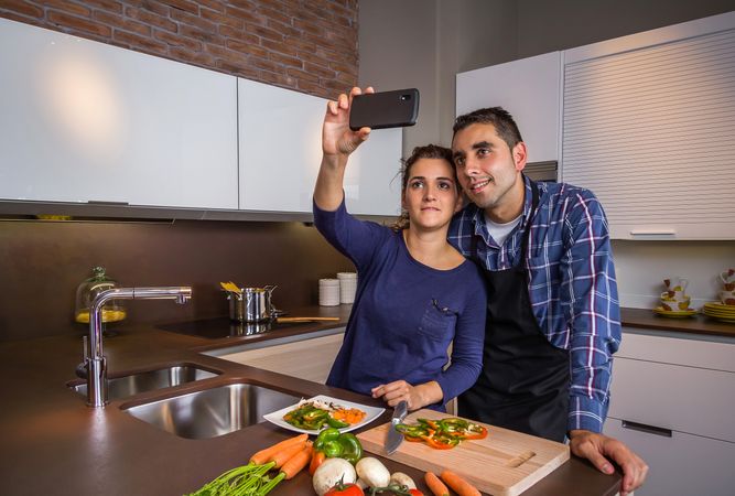 Couple in a home kitchen taking a selfie with a smartphone while preparing meal