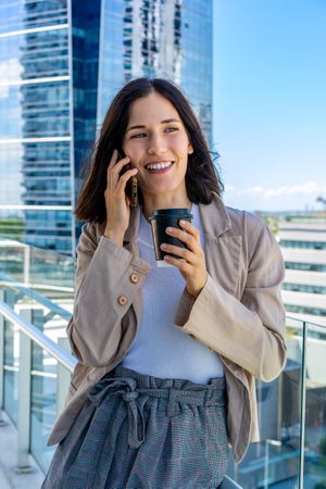 A woman is standing on a balcony and talking on her cell phone on her office break.  She has a paper cup of coffee in her hand.