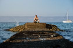 Woman practicing yoga while sitting on boulder near blue ocean