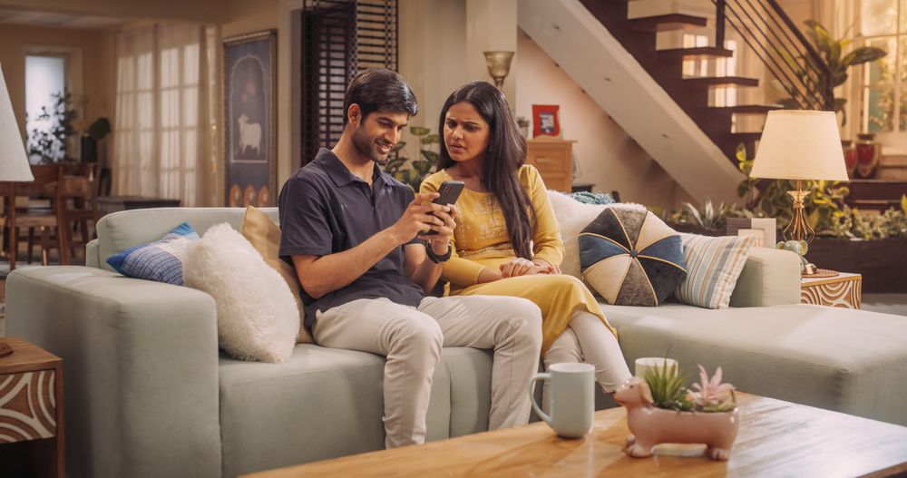Lovely Young Indian Couple Using Internet On Smartphone, Sitting On The Sofa at Home, they Tease and Joke around.