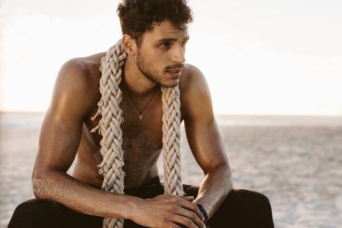 Young man relaxing after intense workout at a beach with a battle rope around his neck
