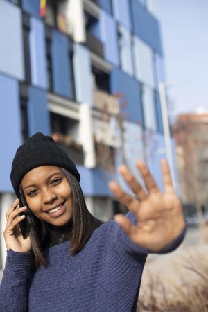 Female in wool hat and blue sweater blocking camera with hand while talking on cell phone