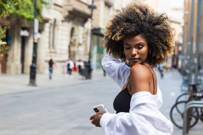 Black woman touching hair while standing on street with smartphone