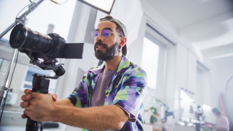Focused Male Cinematographer With Beard, Wearing Colorful Shirt, Operates Professional Camera In Bright Studio Setting, Capturing Engaging Content For Film Or Advertising Project.