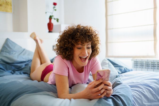 Woman reacting to something on her phone while lying on bed on her stomach