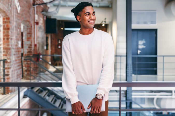 Young stylish man standing in modern office holding a laptop smiling and looking away