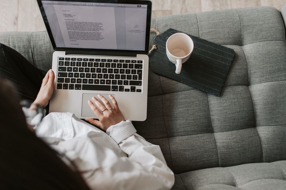 Top view of woman in light shirt using a laptop sitting on gray couch beside empty mug