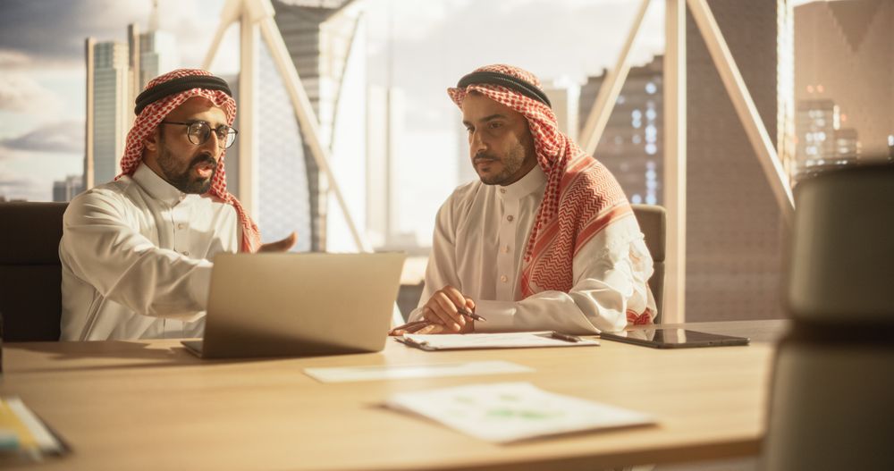 Two Young Muslim Financial Advisers Use Laptop Computer to Discuss a Stock Market Strategy in a Modern Industrial Office.