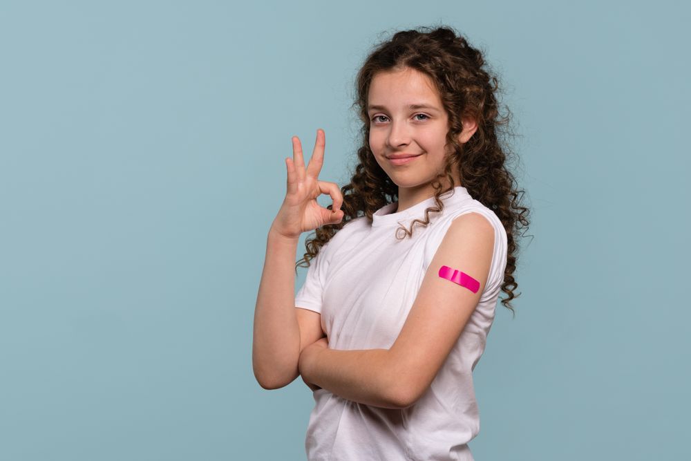 Girl making "OK" gesture with pink bandage on vaccinated arm.