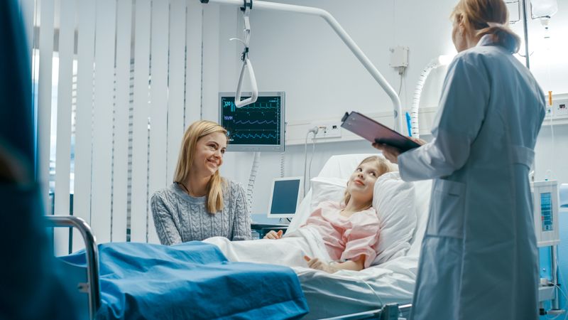 Recovering Little Girl Lies on a Bed In the Hospital, Friendly Doctor With Clipboard Asks Where it Hurts, Mother Sits Beside Bed.