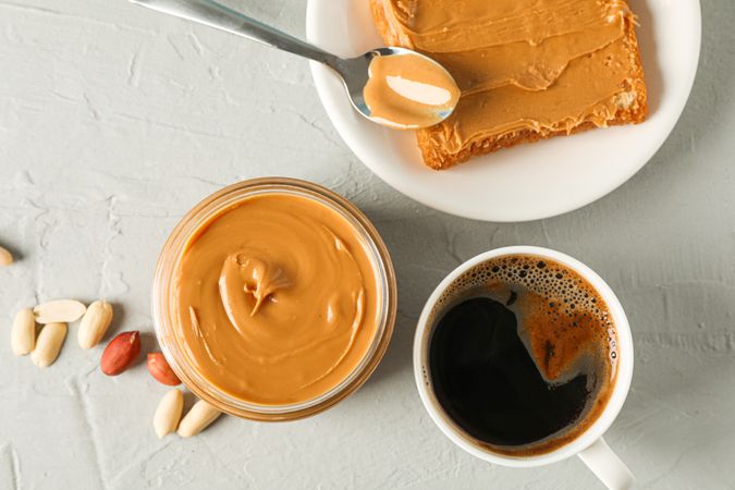 Glass jar with peanut butter, peanut, cup of coffee, spoon and peanut butter sandwich on grey background, top view and space for text