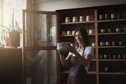 Smiling woman in leather apron inspecting handmade plates in her shop