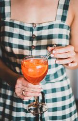 Young woman in checkered dress stirring aperitif with orange slices and glass straw