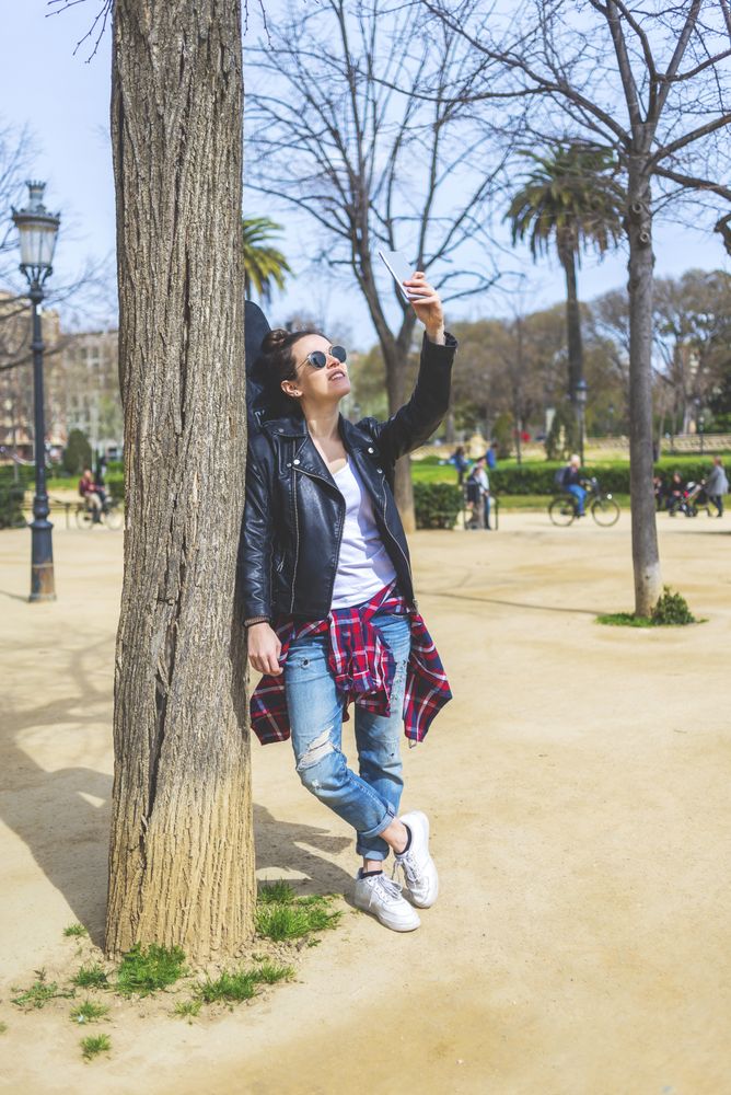 Side view of a young laughing hipster woman standing and leaning on a tree at park in sunny day while using a mobile phone to take a selfie
