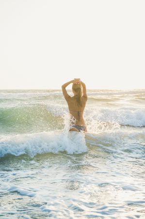 Rear shot of woman in bikini in waves at the beach with hands to her head