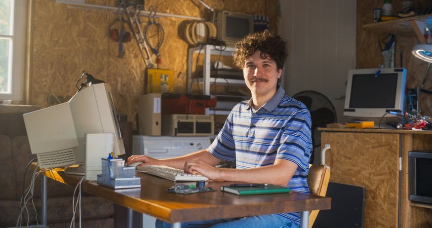 Male Software Engineer Programming On Old Desktop Computer In Retro Garage, Looking At Camera And Smiling.