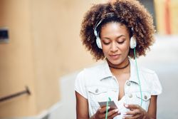 Black female walking down the street wearing headphones and switching songs on phone