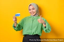 Muslim woman in headscarf and green blouse holding credit card and giving thumbs up 0Lo6yb