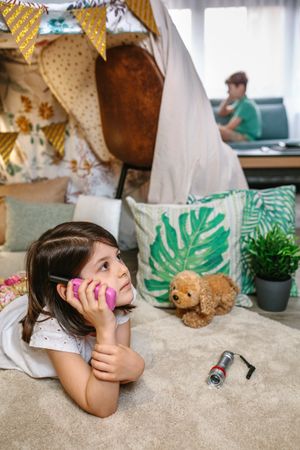 Pensive little girl holding walkie-talkie lying over rug in tent while talk with boy sitting on sofa
