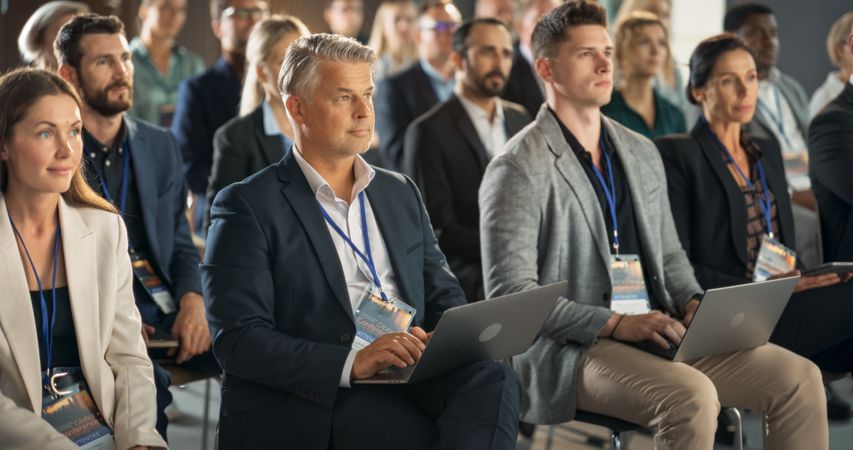 Male Pharmaceutical CEO Sitting In Diverse Crowd At Medical Summit And Using Laptop Computer.