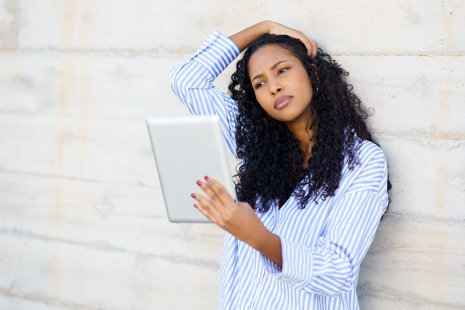 A thoughtful woman engages with a tablet, reflecting on her surroundings against a wall