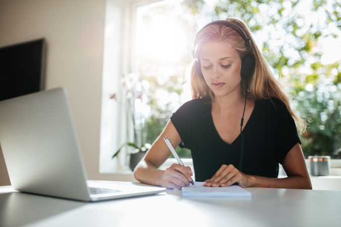 Female wearing headphones and writing with laptop on table at home